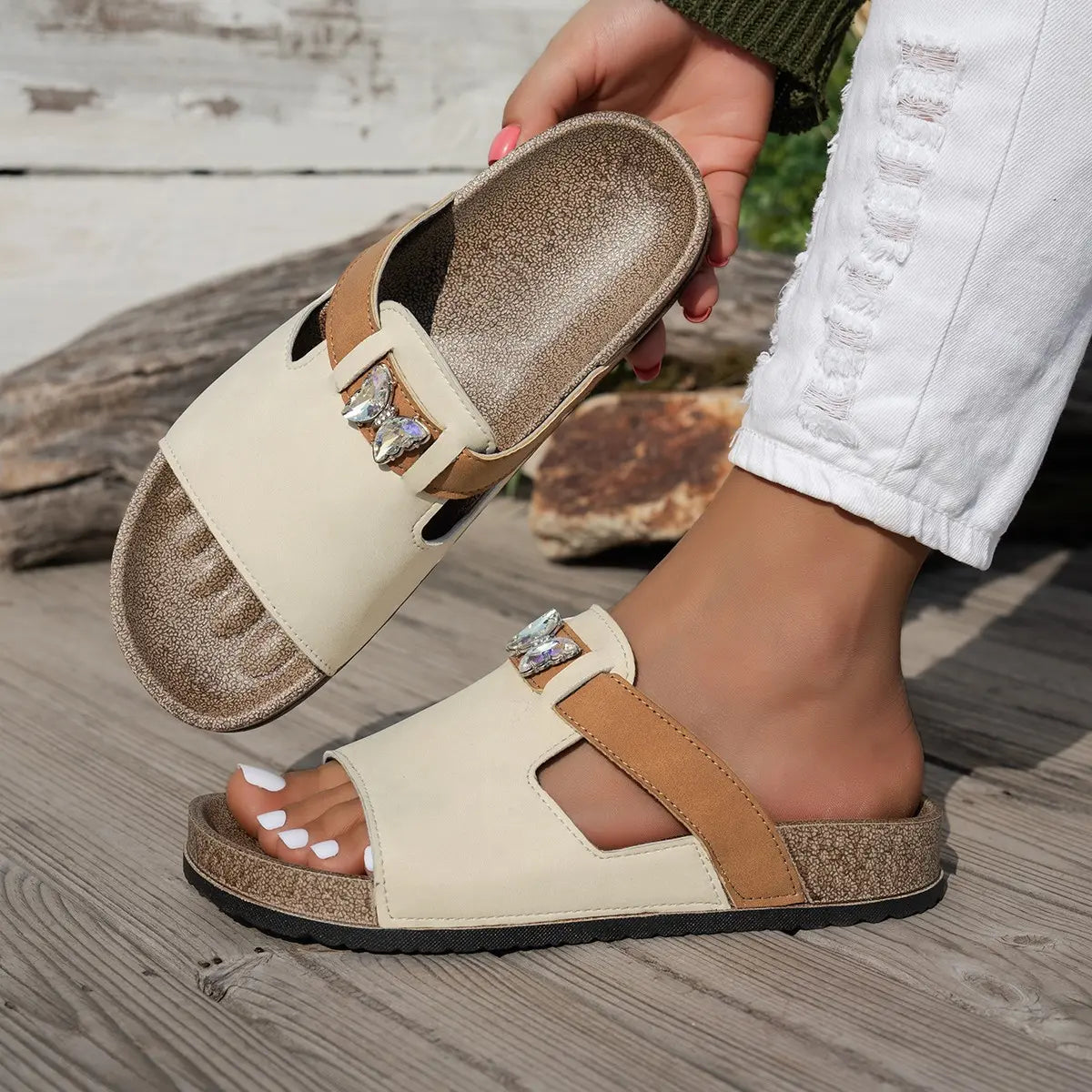 A pair of embellished Xylia flat slide sandals in ivory cream with a tan accent strap, featuring a clear butterfly embellishment, shown worn on a foot outdoors on a wooden deck.