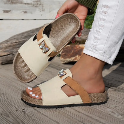 A pair of embellished Xylia flat slide sandals in ivory cream with a tan accent strap, featuring a clear butterfly embellishment, shown worn on a foot outdoors on a wooden deck.