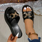 Black Xyloen women's wedge slide sandals featuring a chunky metallic gold chain detail across the peep-toe strap and a textured wedge heel. One sandal is held by a hand, showing the sole, while the other is worn to display the fit.