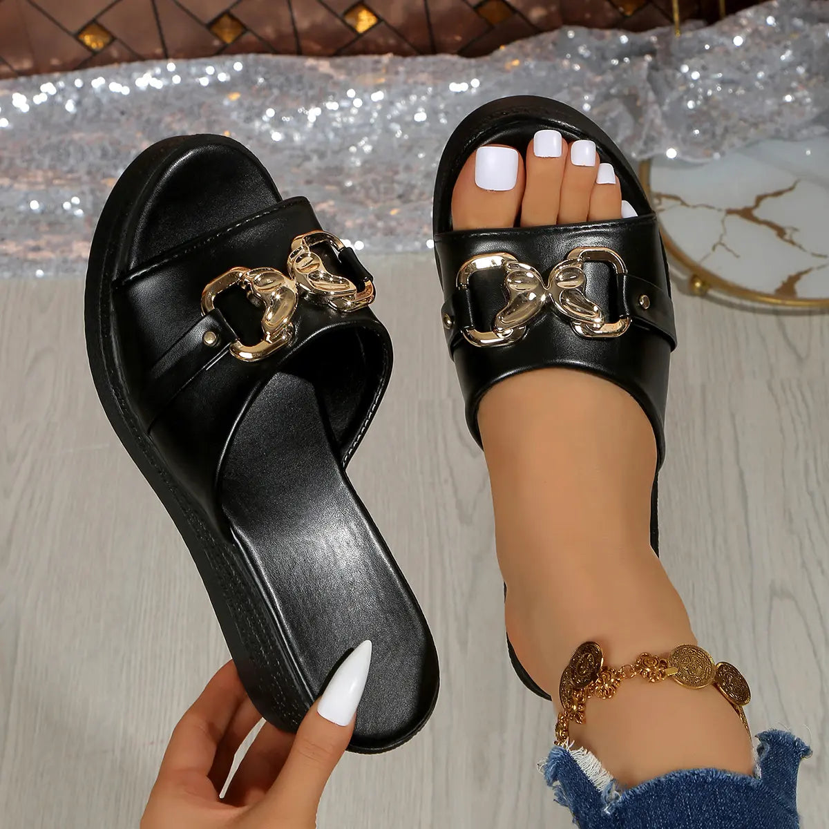 Black Xyloen women's wedge slide sandals featuring a chunky metallic gold chain detail across the peep-toe strap and a textured wedge heel. One sandal is held by a hand, showing the sole, while the other is worn to display the fit.