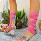 A pair of Xyloq platform wedge sandals adorned with a vibrant, glossy psychedelic swirl print and hot pink elastic gladiator straps wrapping up the leg, shown on feet in an outdoor setting with cacti and decorative elements.