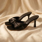 Black Xylos square-toe stiletto mules with a knot detail across the vamp, shown from a front angle on a draped fabric background.