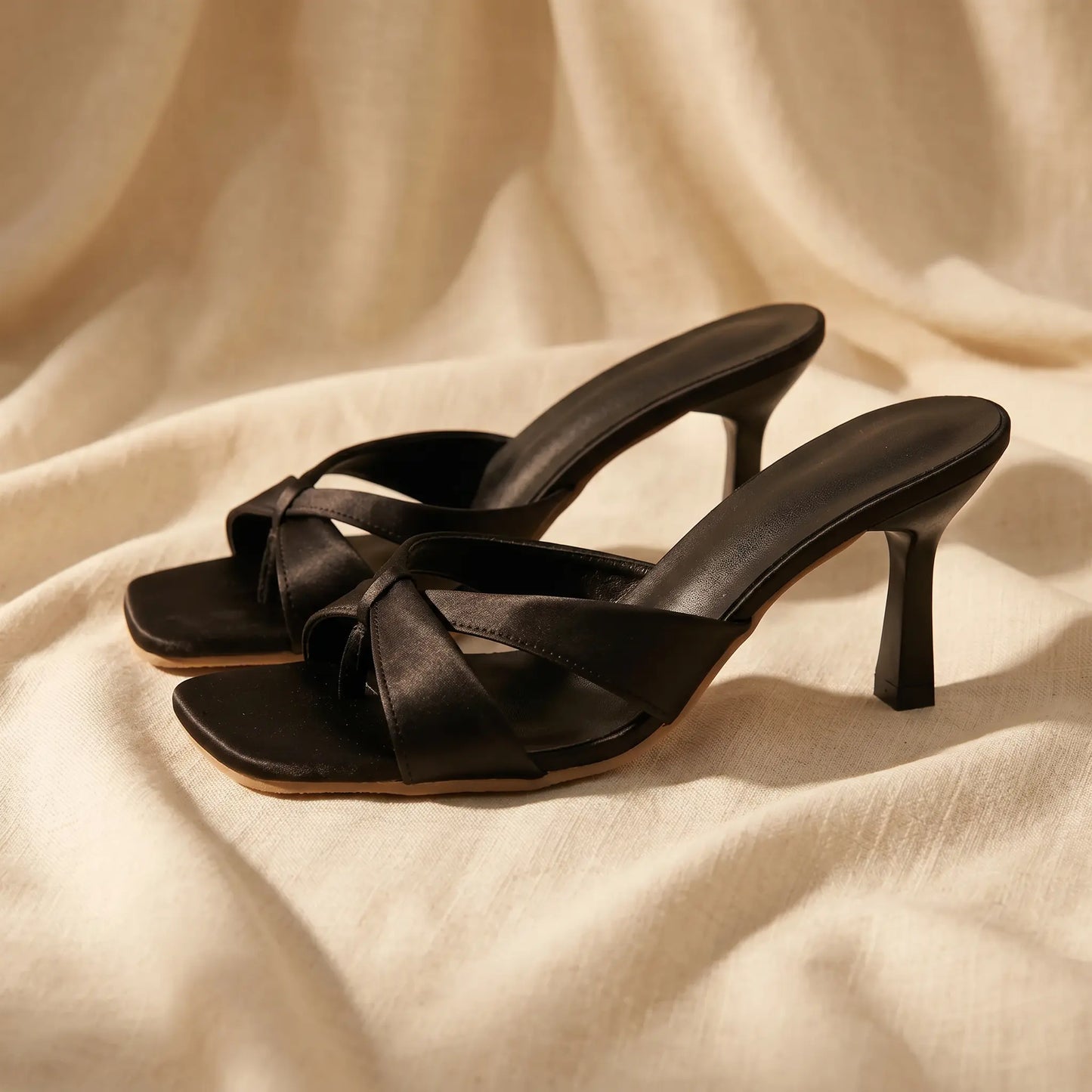 Black Xylos square-toe stiletto mules with a knot detail across the vamp, shown from a front angle on a draped fabric background.