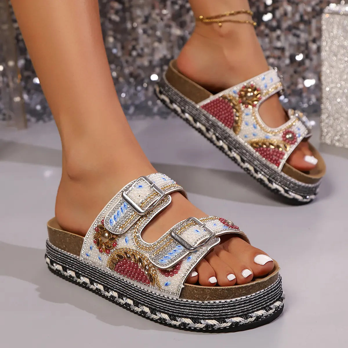 Embellished platform slide sandals with double buckle straps and intricate beadwork shown on a model's feet against a shimmering studio background.