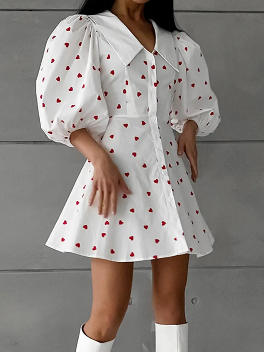 White cotton-linen blend midi dress featuring a playful red heart print, dramatic puff sleeves, a pointed collar, and a button-down front.