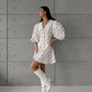 Woman wearing a white Xylos heart print puff sleeve midi dress with a pointed collar and A-line silhouette, styled with white boots against a textured grey wall.