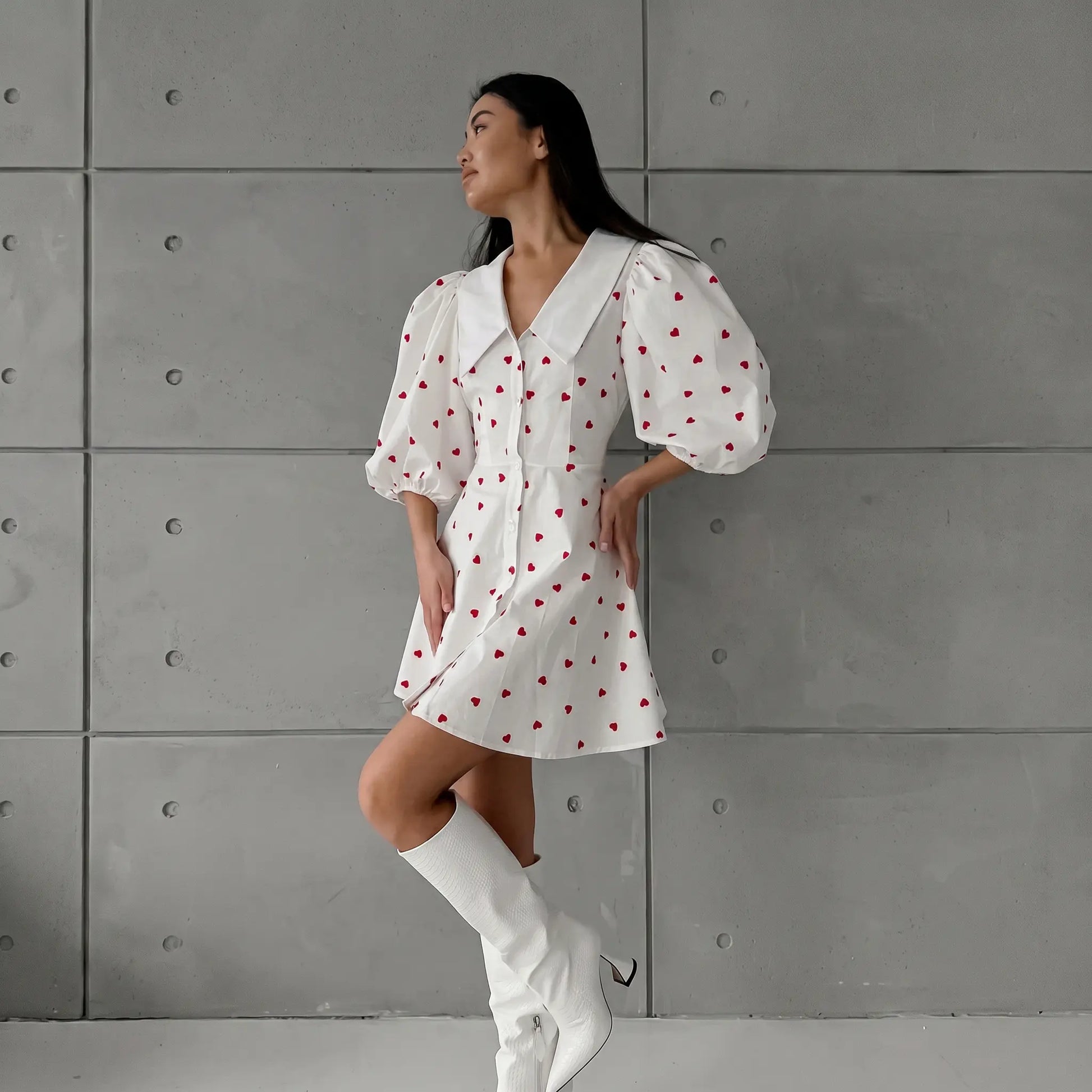 Woman wearing a white Xylos heart print puff sleeve midi dress with a pointed collar and A-line silhouette, styled with white boots against a textured grey wall.