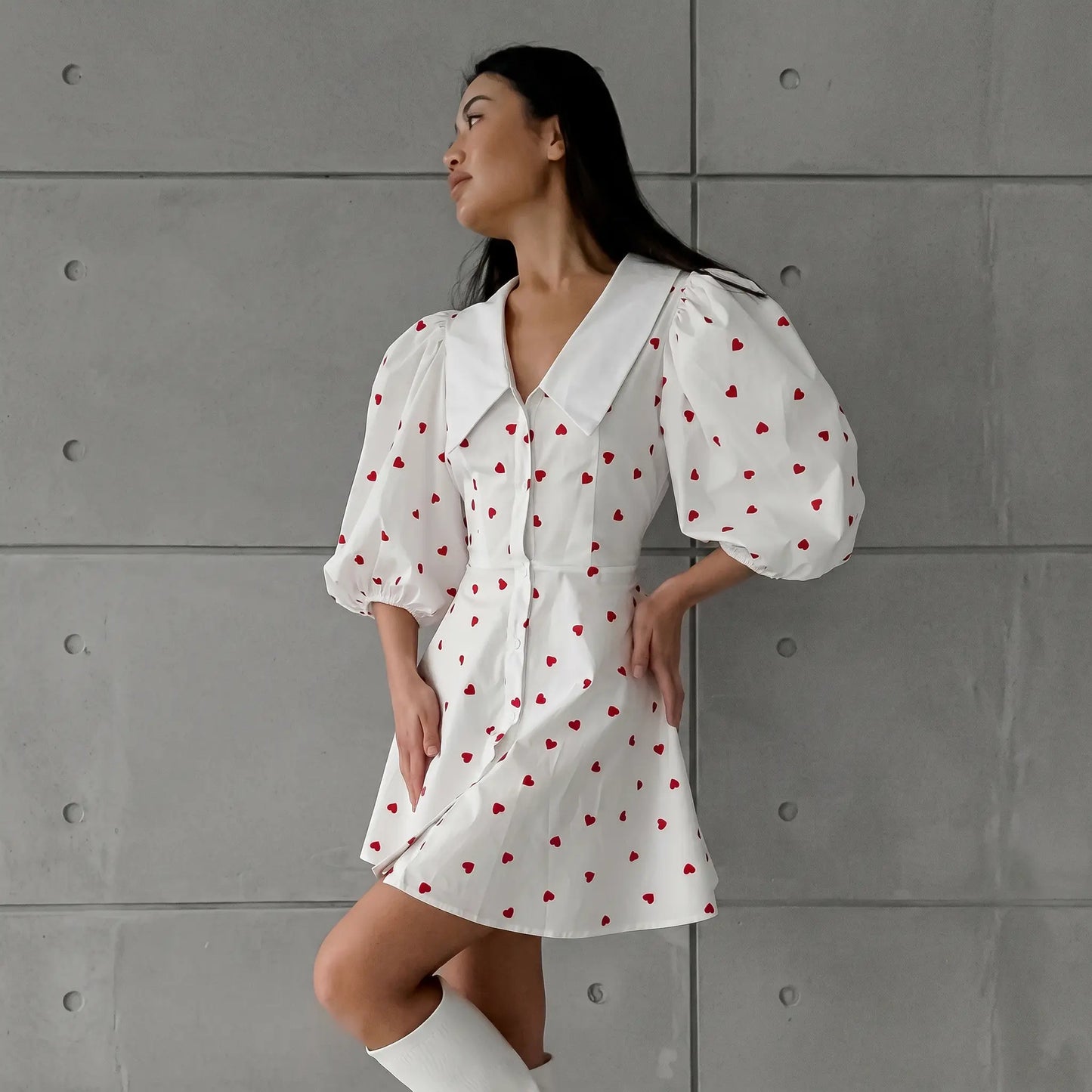 A woman models a white Xylos heart print puff sleeve midi dress with a pointed collar and button-down front, standing against a grey textured wall.