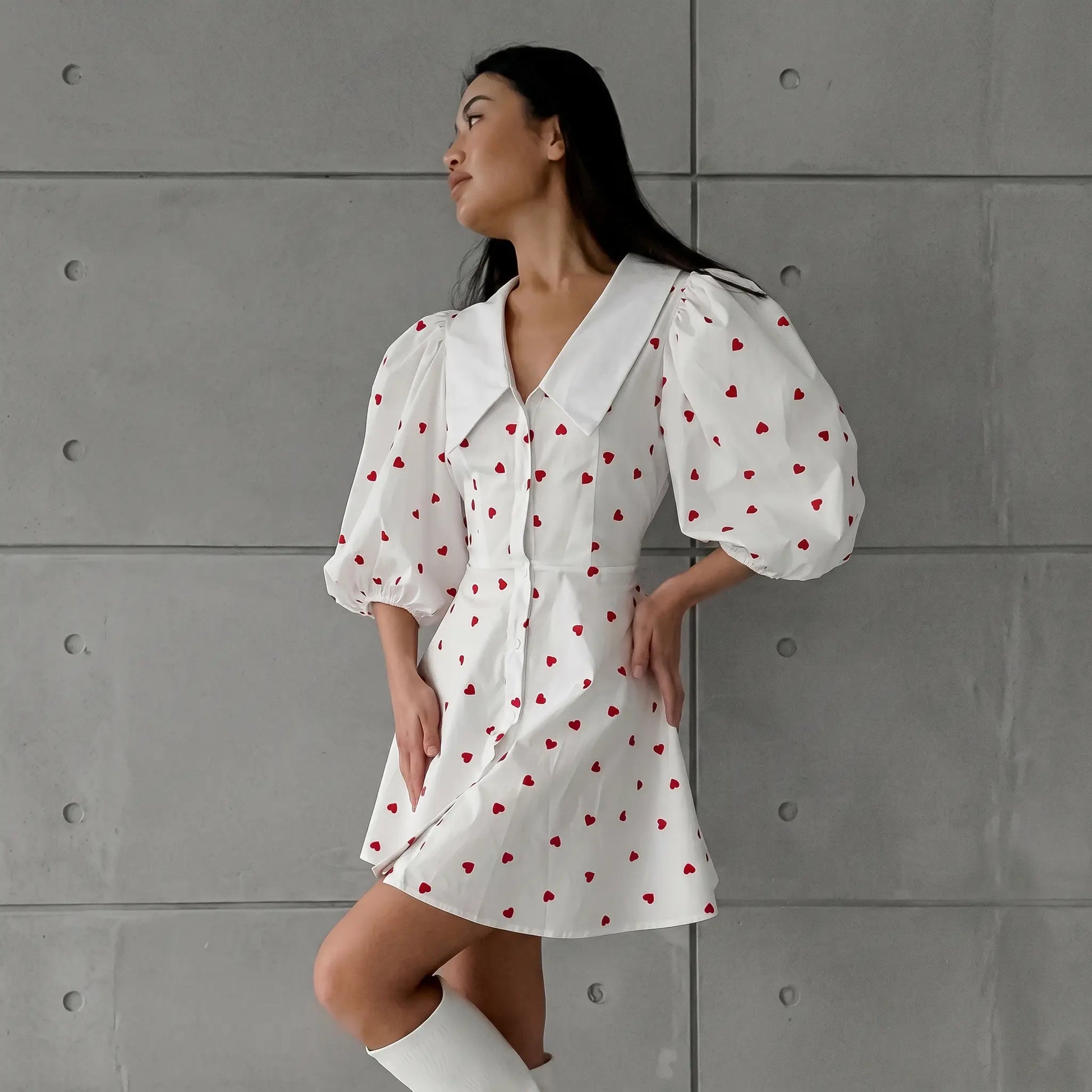 A woman models a white Xylos heart print puff sleeve midi dress with a pointed collar and button-down front, standing against a grey textured wall.