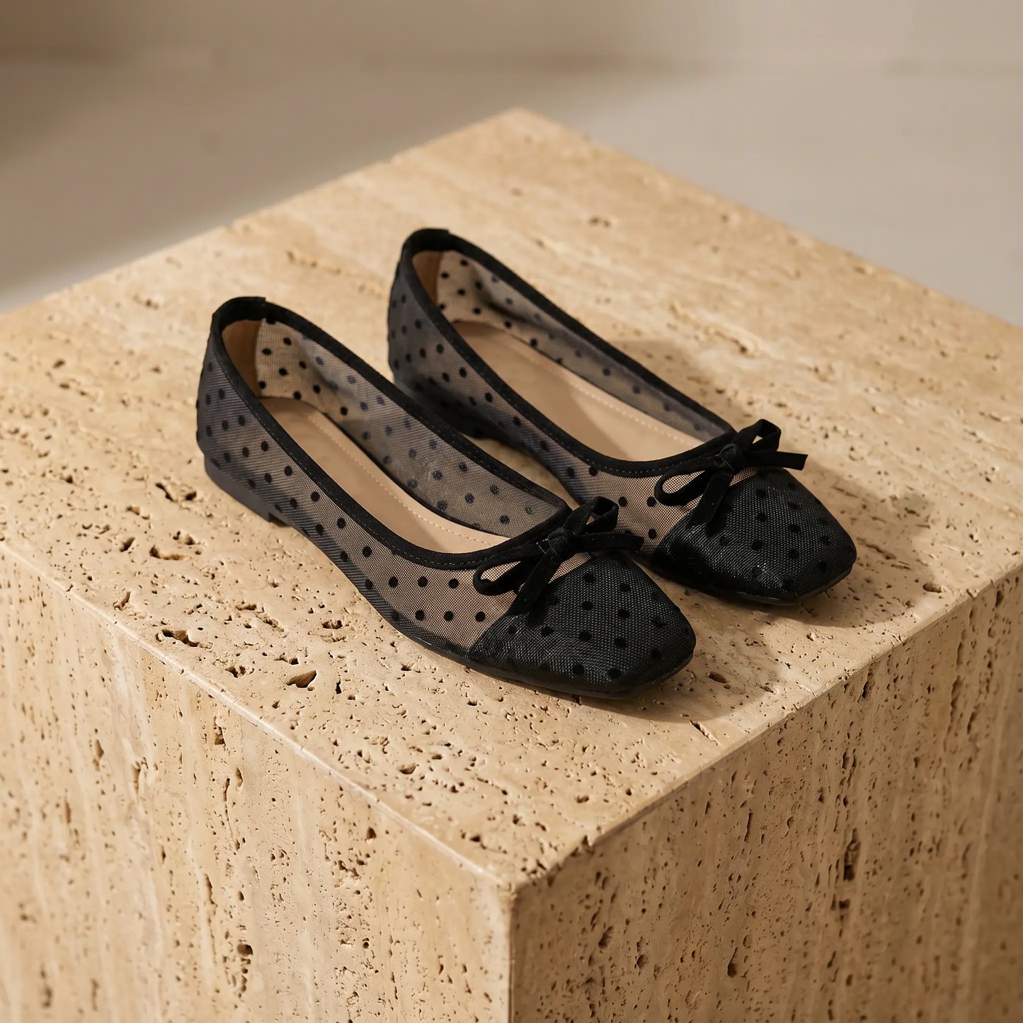 A pair of Xyndra black sheer mesh ballet flats with flocked polka dots, solid toe caps, and delicate ribbon bows, presented on a light brown textured stone pedestal.