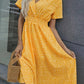 Model wears a vibrant yellow ditsy floral print midi dress with a V-neck, flutter sleeves, and a smocked waist, holding a wooden bag.