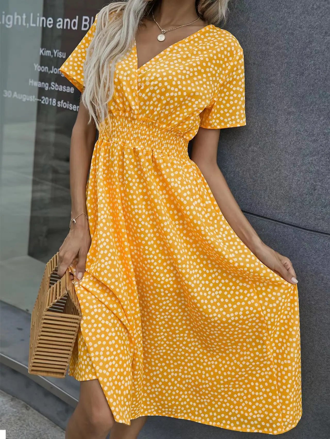 Model wears a vibrant yellow ditsy floral print midi dress with a V-neck, flutter sleeves, and a smocked waist, holding a wooden bag.
