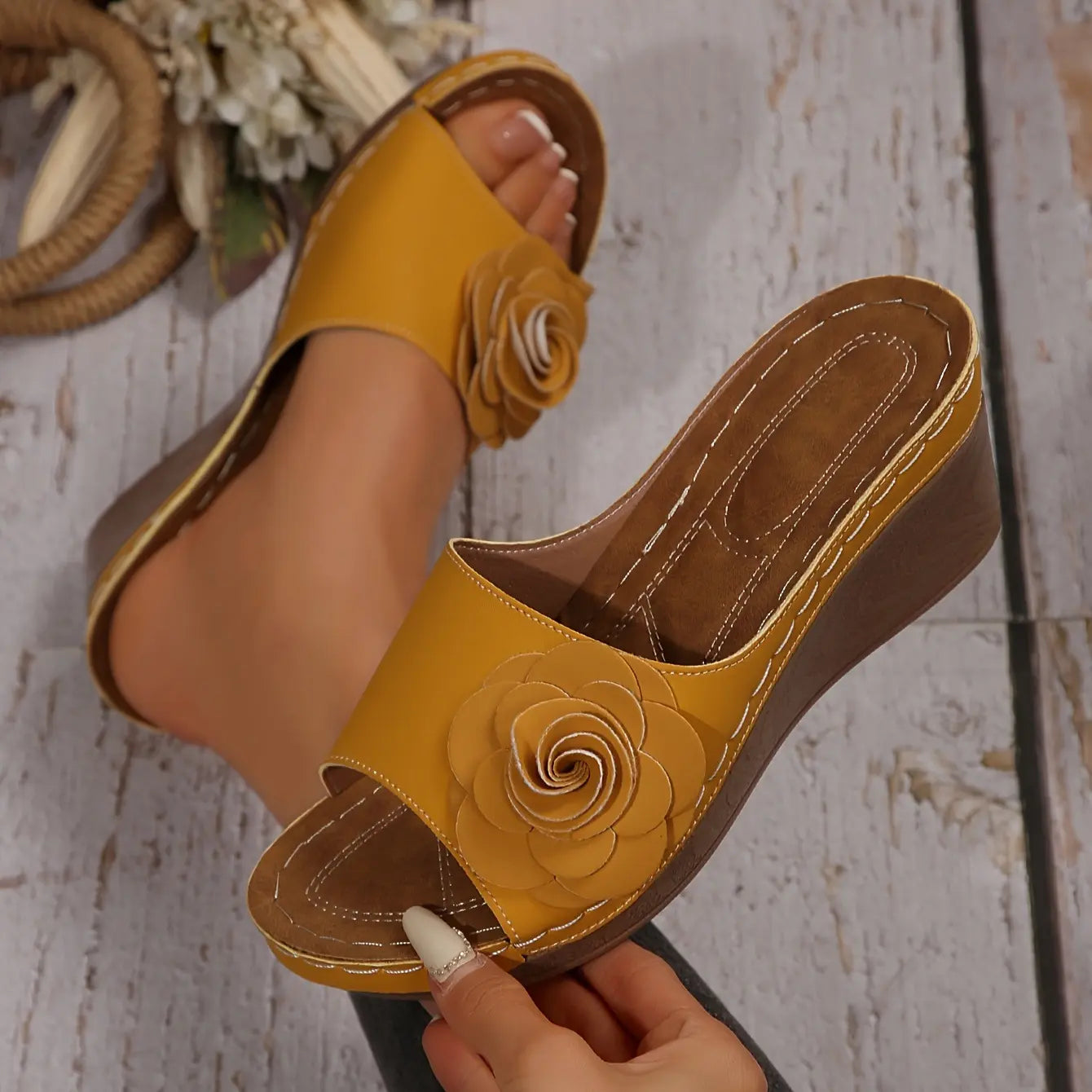 Women's yellow slip-on wedge sandals featuring a large fabric rose embellishment and white contrasting stitching, shown on a foot in a lifestyle setting with a rustic wooden background.