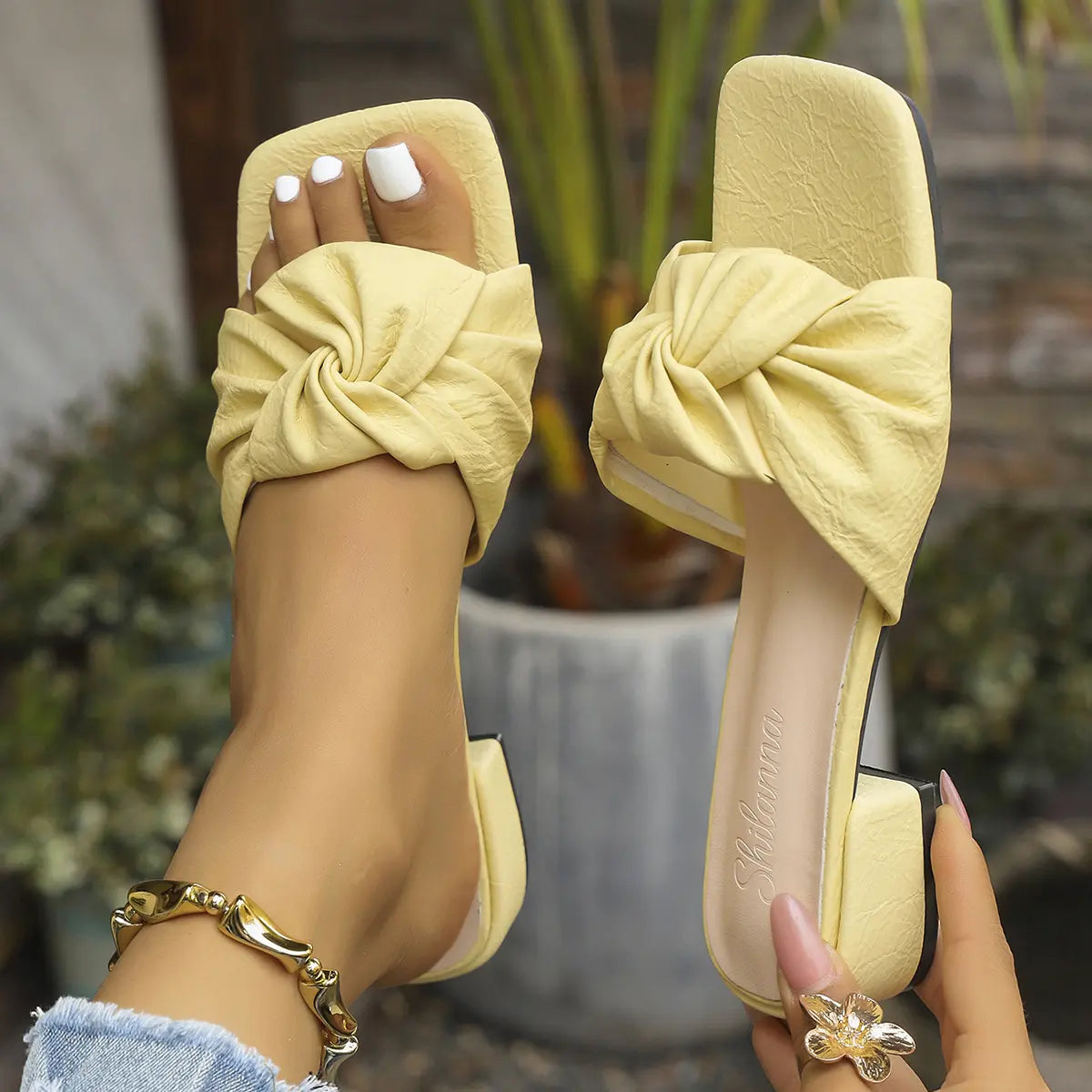 Elegant yellow low-heel slide sandals with a chic knot strap, shown on a woman's foot adorned with a gold bracelet, showcasing square toe and comfortable block heel.