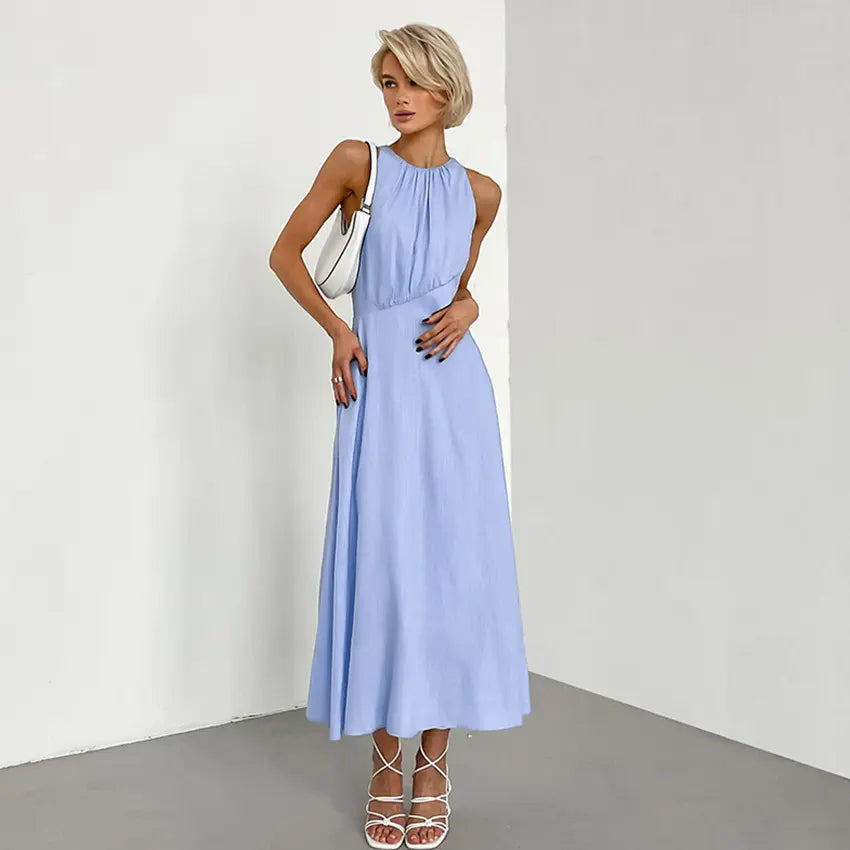 Sleeveless pale blue Zarion midi dress with a gathered halter neckline and side waist cut-outs, worn by a model with a white handbag and white sandals against a studio background. This romantic bohemian dress showcases a flowy A-line skirt and linen-blend fabric.