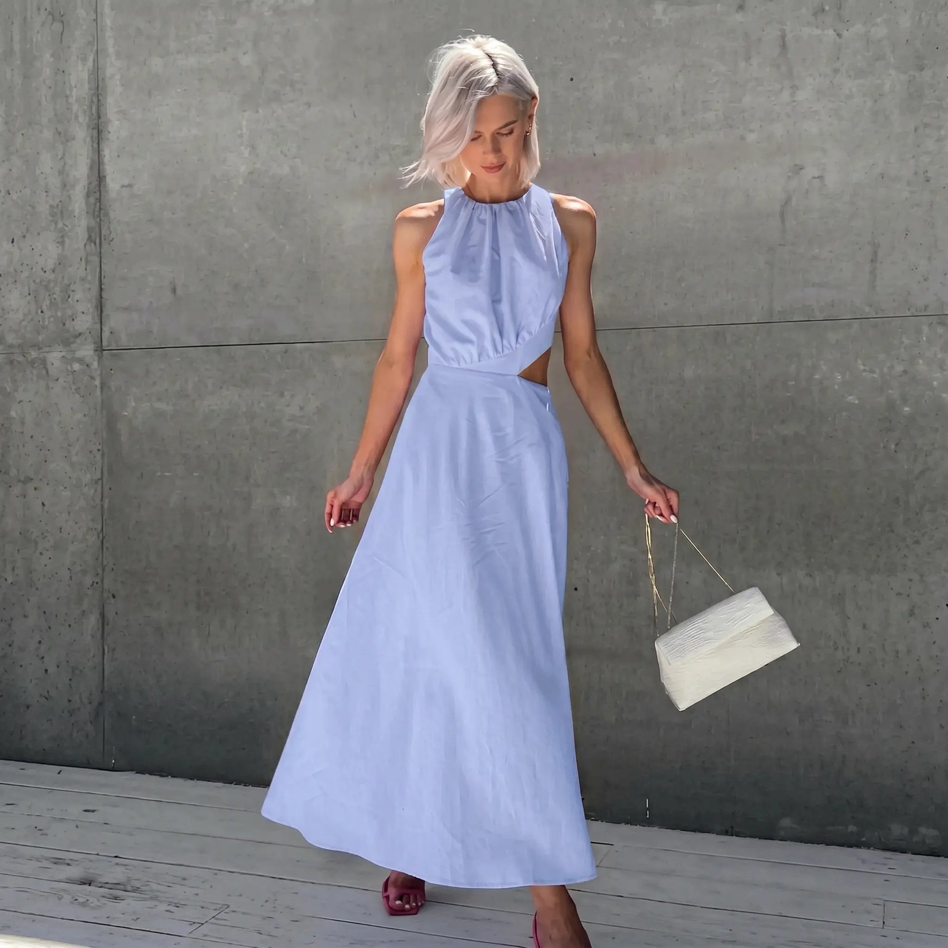 A romantic Zarion sleeveless midi dress in Periwinkle Sky, featuring a gathered halter neckline and dramatic side waist cut-outs, shown on a model against a grey wall.