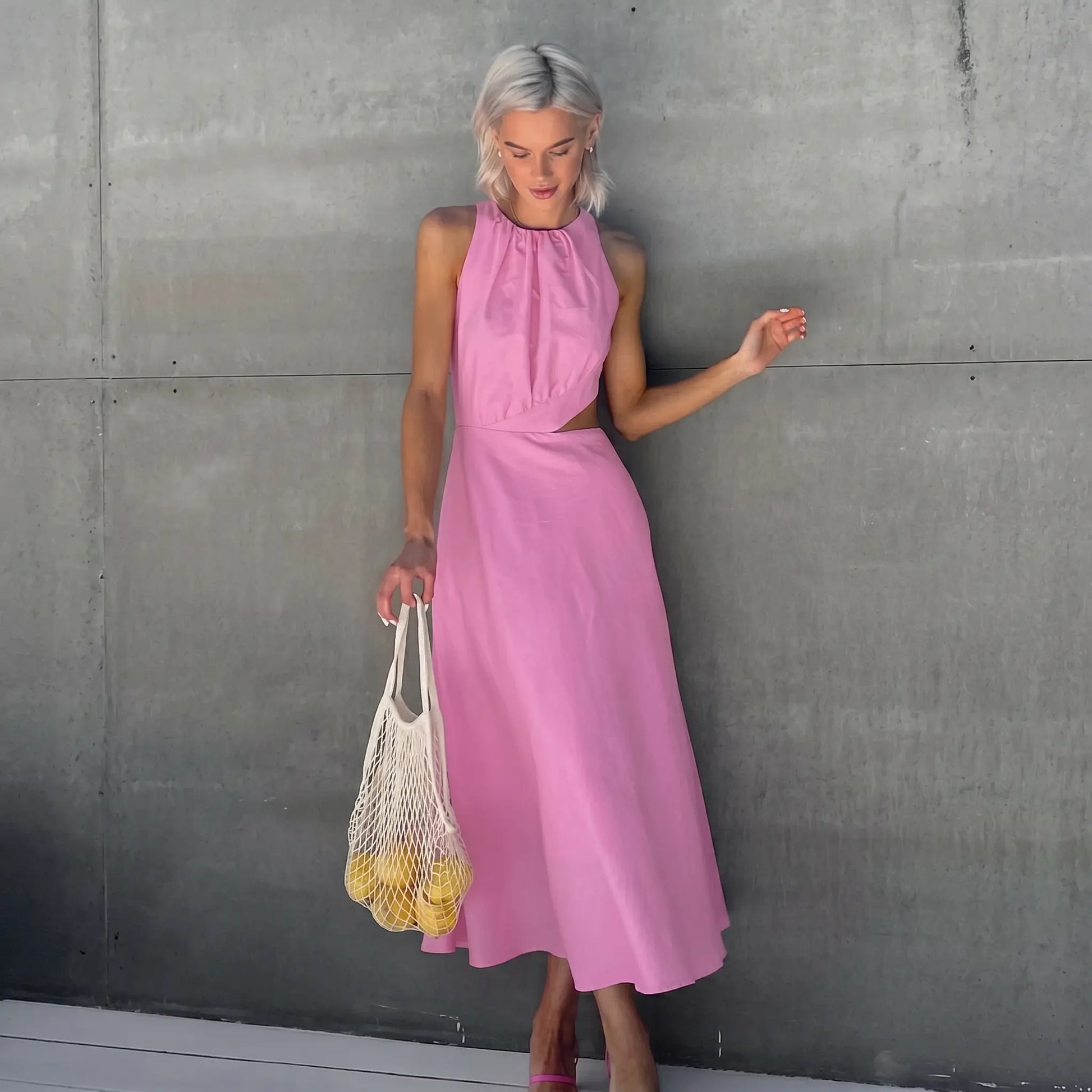 Model wearing a pink sleeveless gathered halter neck midi dress with elegant side waist cut-outs, holding a mesh bag of lemons.