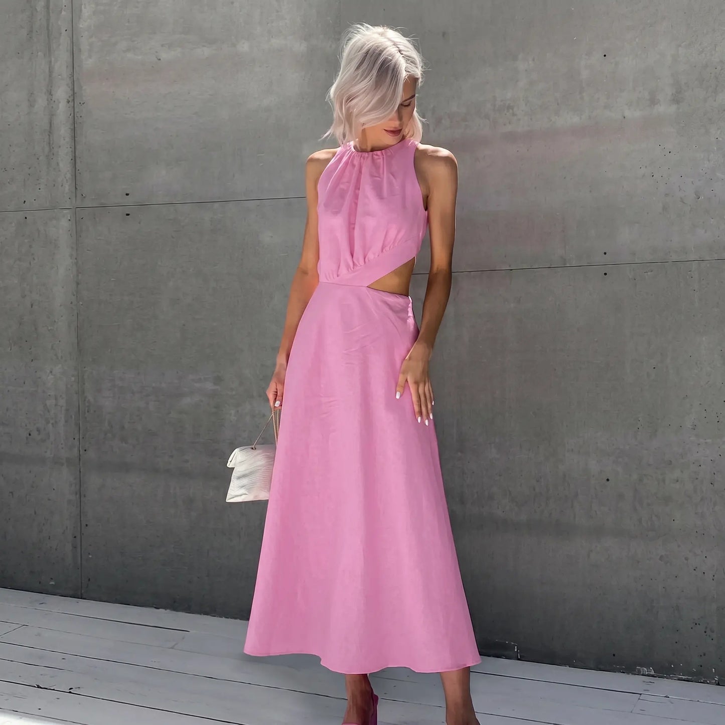 A woman models a sleeveless Zarion pink midi dress with romantic gathered halter neckline and dramatic side waist cut-outs, set against a textured grey wall.