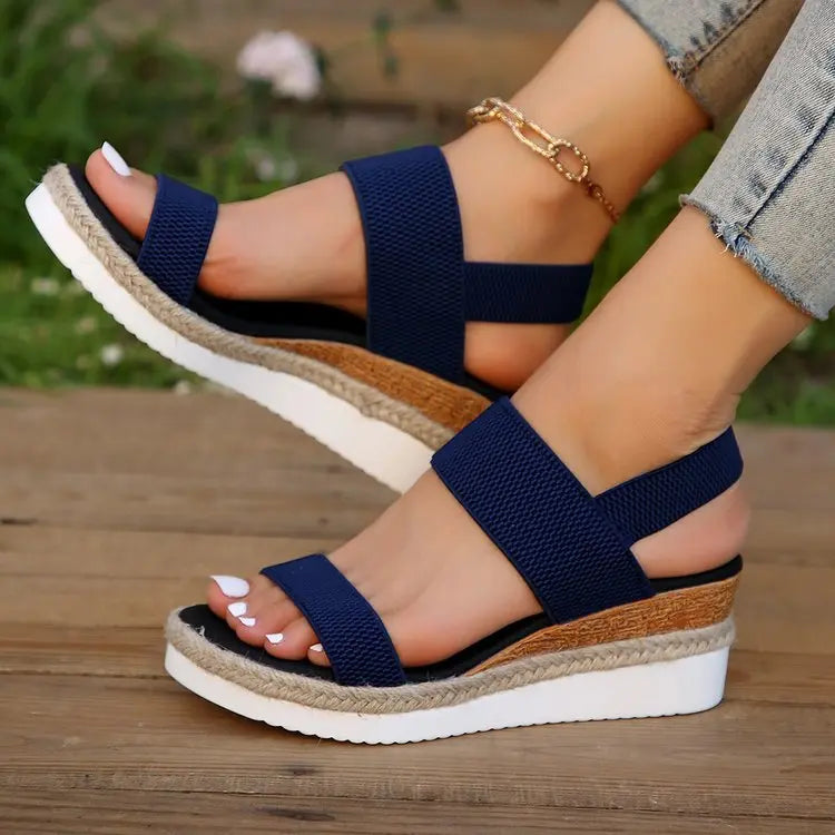 Navy blue Zaylin espadrille wedge sandals with peep-toe and elastic straps, worn by a model with a gold-tone ankle chain on a wooden deck with greenery in the background.