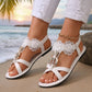 A pair of Zaylor women's white flat sandals, adorned with intricate floral lace appliques, pearls, and a sparkling rhinestone oval buckle, worn on a sandy beach at sunset with gentle waves in the background. A gold anklet is visible.