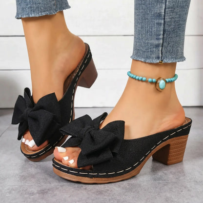 Black Zayna heeled mule sandals featuring a large knotted bow, peep-toe design, chunky block heel, and white contrast stitching, worn with denim jeans.