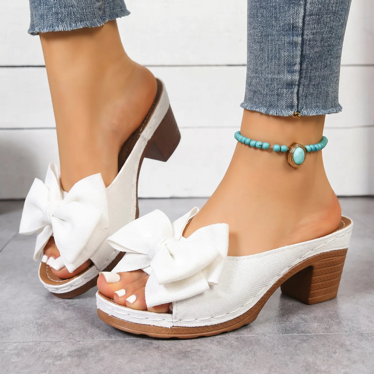 Zayna white heeled mule sandals with oversized knotted bow detail, peep-toe, and chunky block heel, shown on a model's feet wearing denim jeans.