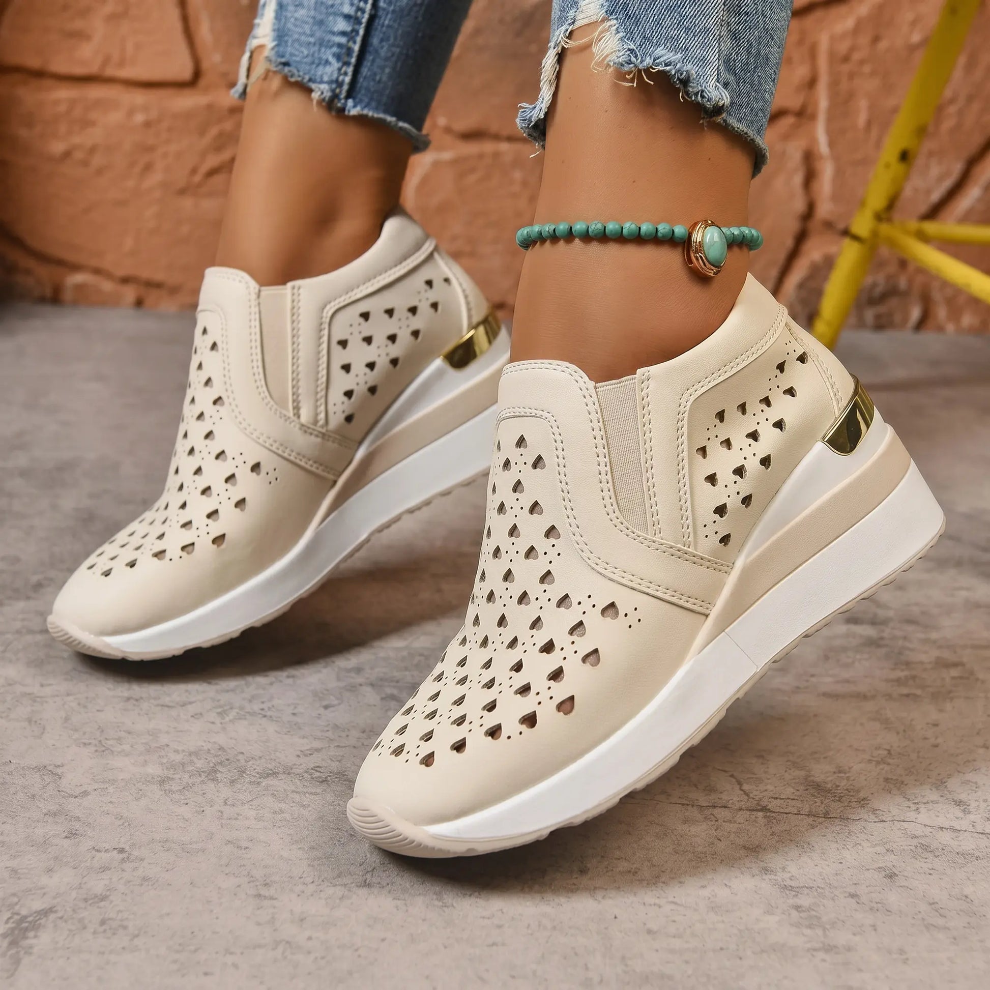 Women's beige perforated slip-on loafers with a hidden wedge heel, worn with ripped jeans and a turquoise anklet. The shoes feature a heart-shaped cutout pattern and a gold heel accent.