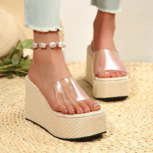 Women's Zelka clear strap wedge slide sandals in almond beige, worn with distressed jeans and a pearl anklet on a woven mat.