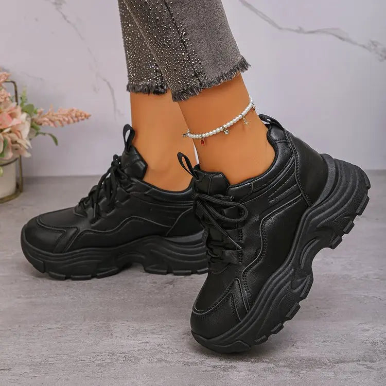 A pair of black Zelpha chunky platform sneakers with a synthetic leather upper and lugged rubber sole, worn on feet with embellished grey denim jeans and a pearl anklet.