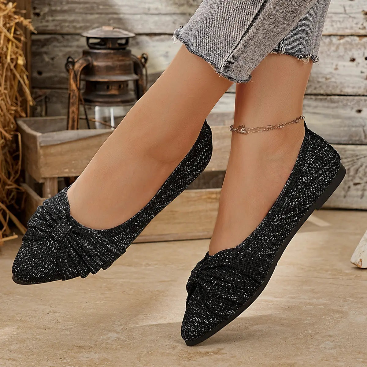 Close-up of elegant black knit pointed-toe flats with a bow detail, worn by a model with ripped jeans and an anklet, showcased in a rustic lifestyle setting.