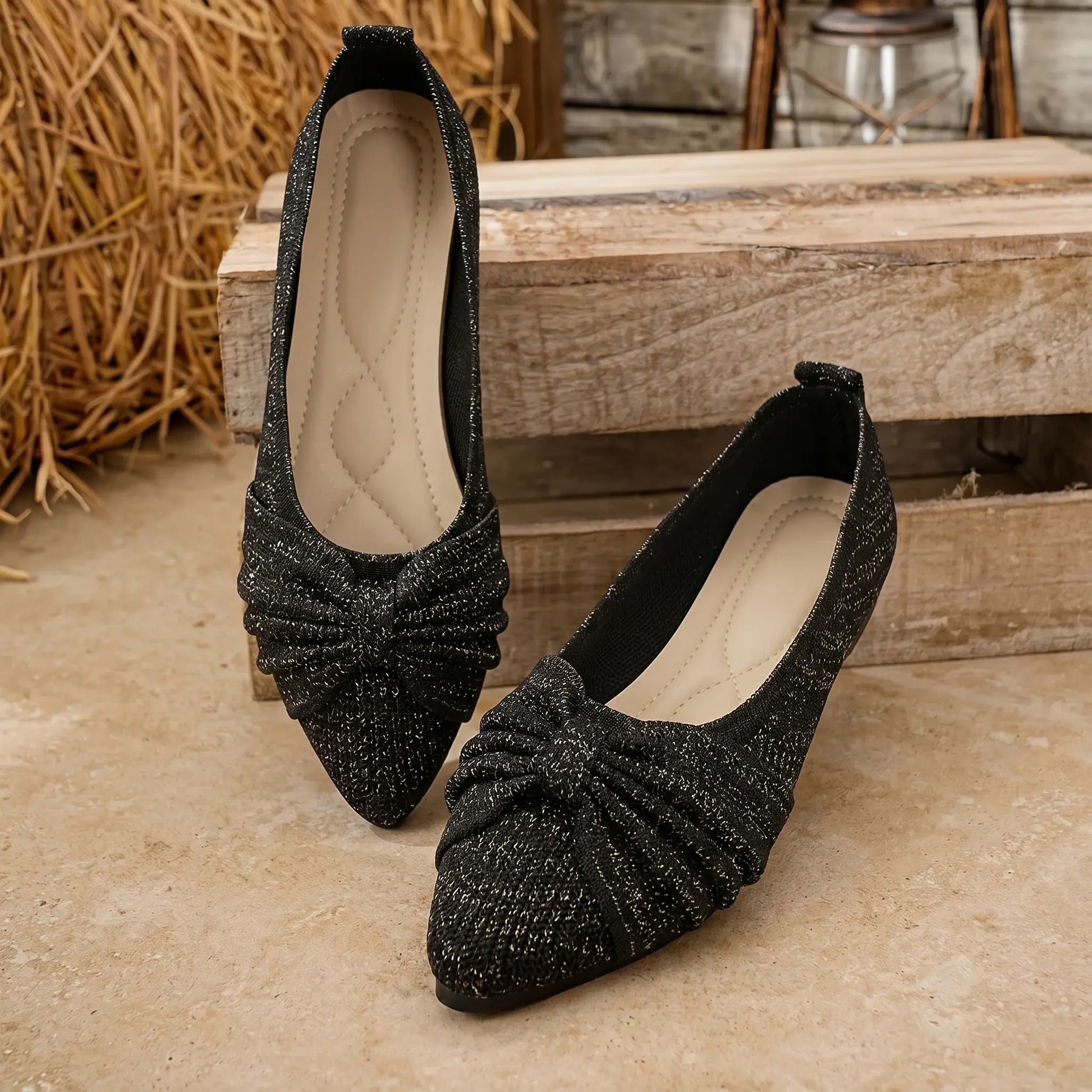 A pair of Zendari women's midnight black knit slip-on flats with a pointed toe and a prominent bow detail, featuring a subtle metallic shimmer and a flat sole, presented in a rustic lifestyle setting.