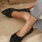Elegant black knit flats for women with a pointed toe and delicate bow detail, shown on a model wearing grey jeans, styled with an anklet. These comfortable chic footwear pieces feature a subtle metallic shimmer for sophisticated day-to-night wear.