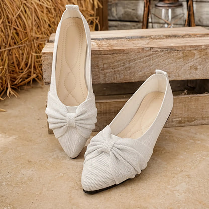 A pair of white Zendari women's slip-on flats featuring a pointed toe and a prominent bow accent, displayed on a tiled surface with rustic wooden crates and straw in the background.
