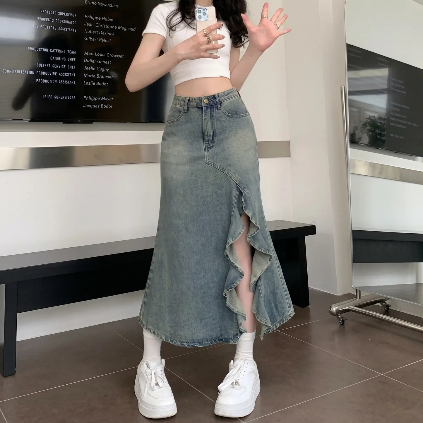 Model wearing a Zendru vintage blue high-waisted asymmetrical ruffled denim midi skirt. The skirt features a dramatic diagonal ruffled slit, revealing the leg and styled with a white crop top and sneakers.