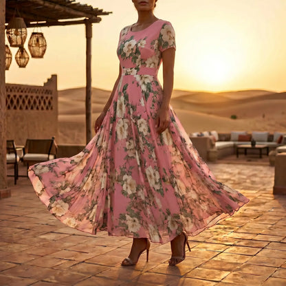 Woman in Zendyr Blush Pink Floral Chiffon Midi Dress, featuring a scoop neck, short sleeves, and a flowing A-line skirt with a wide waistband. Model stands elegantly on a terracotta patio against a desert sunset backdrop, showcasing the delicate watercolor floral print and ethereal drape of the dress.