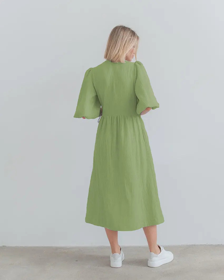 Back view of a flowing midi A-line wrap dress in sage green cotton gauze with voluminous short puff sleeves. The dress features an elasticated waistband and is paired with white sneakers.