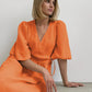 Model wearing a vibrant orange cotton gauze midi wrap dress with short puff sleeves and a tie waist, seated against a neutral background.