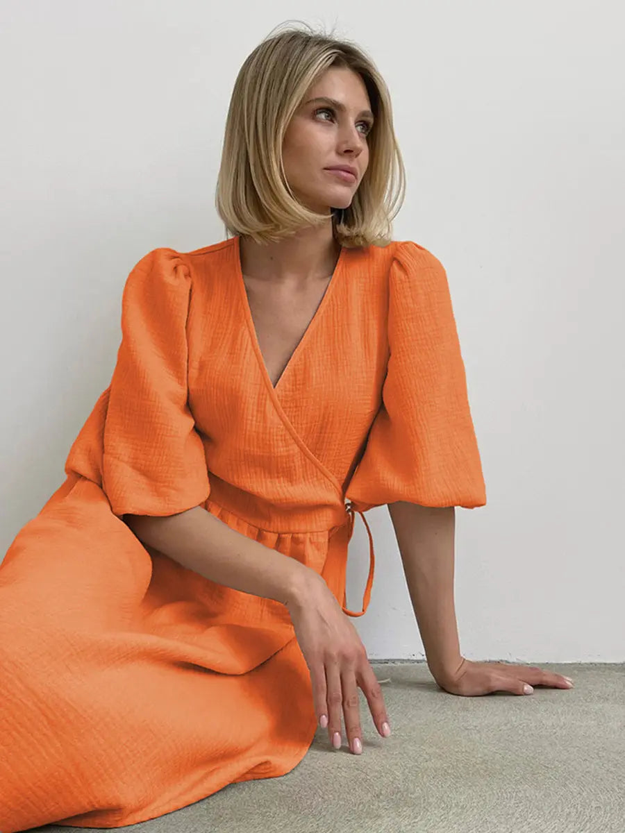 Model wearing a vibrant orange cotton gauze midi wrap dress with short puff sleeves and a tie waist, seated against a neutral background.