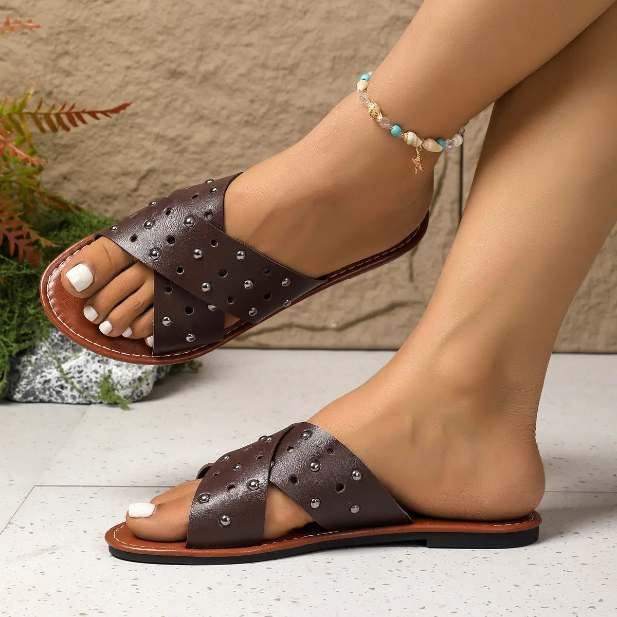 Zenoria studded slide sandals for women, featuring dark brown criss-cross straps with perforations and silver studs, worn on a model's feet with white-painted toenails and a beaded anklet.