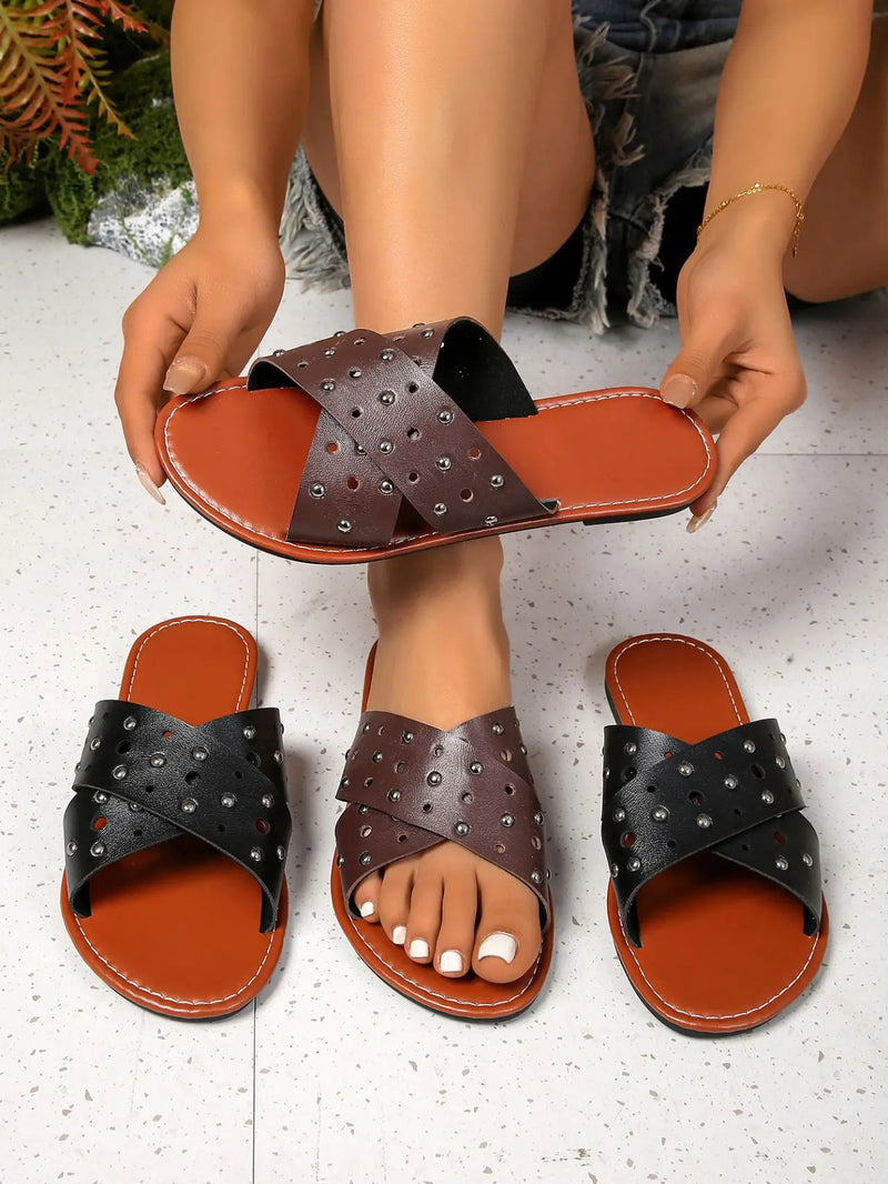 Zenoria studded slide sandals in black and brown with tan footbeds, featuring criss-cross perforated straps and silver-tone studs, displayed on a speckled studio floor with hands holding one pair.