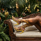 Model wearing vibrant yellow Zephrik lace-up stiletto sandals with slim crisscross straps and extra-long ties wrapped around her calves, reclining on a carved wooden bench in a lush botanical garden.