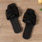 A pair of Zephyra black crushed satin bow square-toe flat slide sandals shown from a top-down view on a light wooden surface with decorative rose petals.