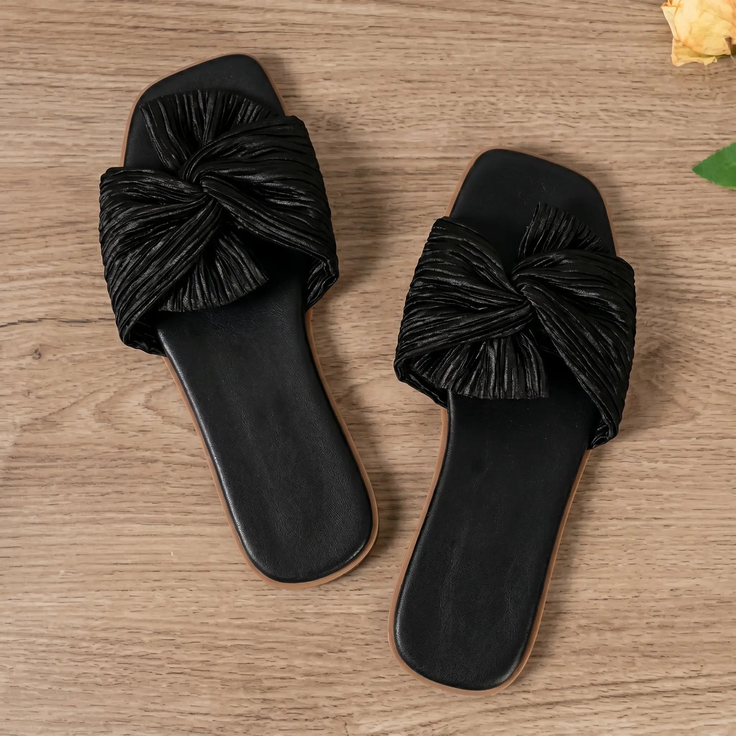 A pair of Zephyra black crushed satin bow square-toe flat slide sandals shown from a top-down view on a light wooden surface with decorative rose petals.