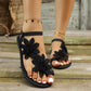 Black Zephyra floral lace flat sandals worn on feet, showcasing delicate appliques, adjustable ankle straps, and a rustic wooden plank background, perfect for summer.