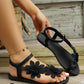 Black Zephyra floral lace flat sandals worn on a woman's feet, showcasing the delicate appliques and adjustable ankle strap against a rustic wooden background. Features white toenails and a subtle gold anklet.