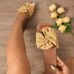 Women's Zephyra crushed satin bow slides in champagne gold, shown on a model's feet with a square toe and flat sole on a wooden surface, accented by yellow roses.