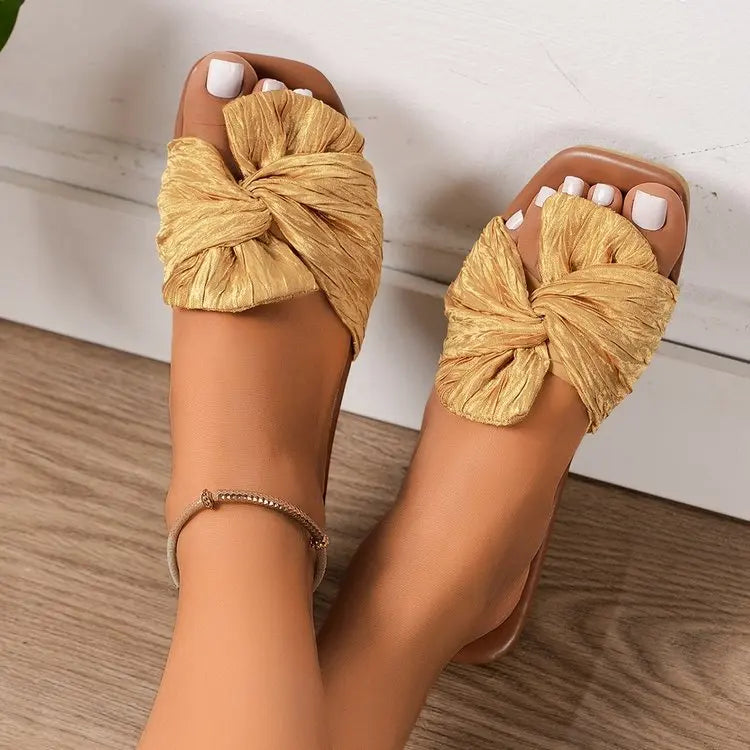 Champagne gold Zephyra women's flat slide sandals featuring a crushed satin bow and square toe, worn on feet against a wooden floor. A delicate anklet is also visible.