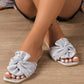 Sterling silver crushed satin bow slides on a model's feet, featuring a square toe and flat sole, shown resting on a wooden floor.