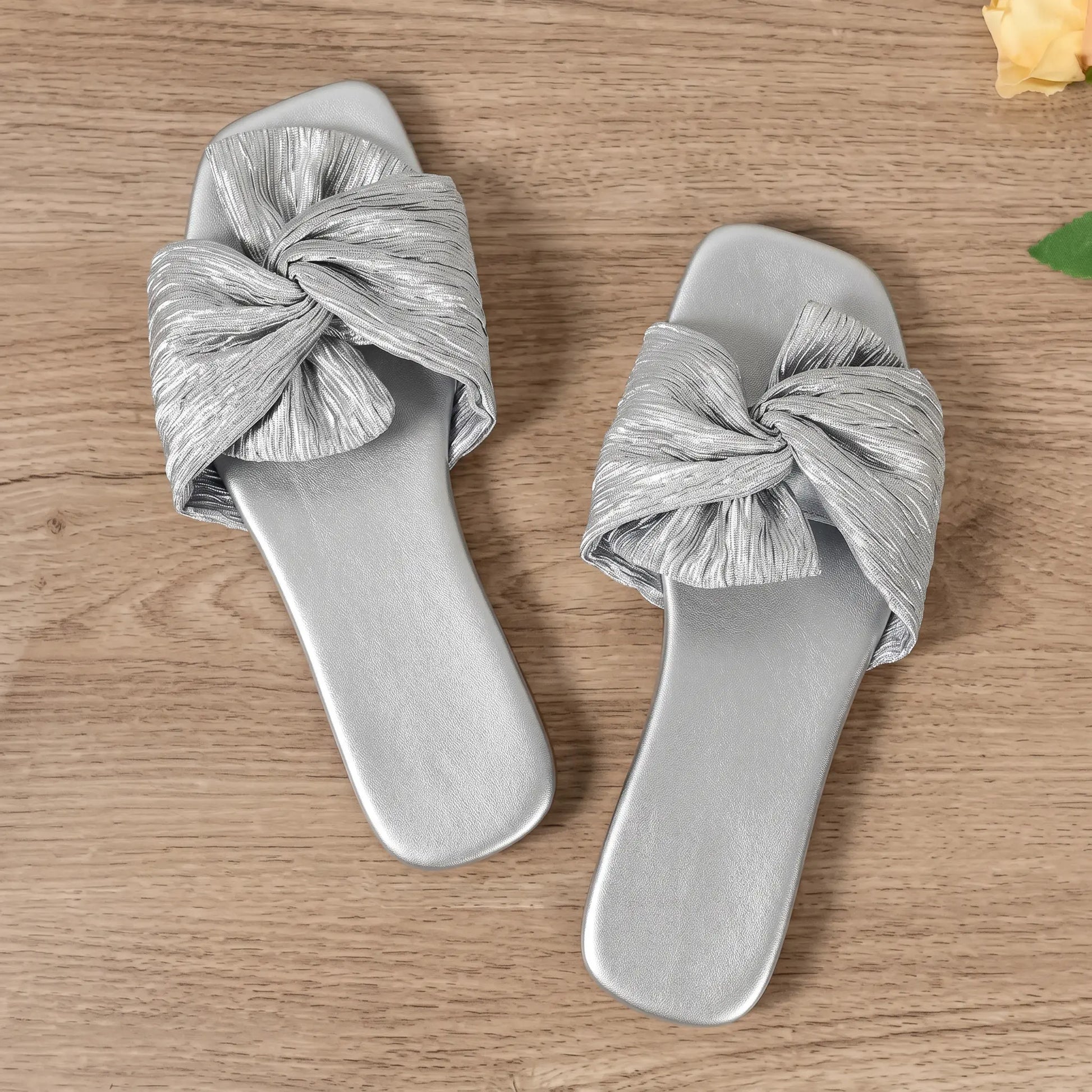 Pair of Zephyra Sterling Silver crushed satin bow square-toe flat slide sandals, viewed from top-down on a wooden surface.