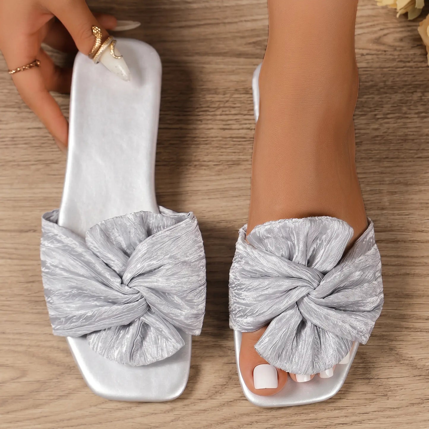 Pair of Zephyra crushed satin bow slides in sterling silver, featuring a square toe and a large knotted bow embellishment, worn on a foot with a second sandal held by a hand against a wooden background.